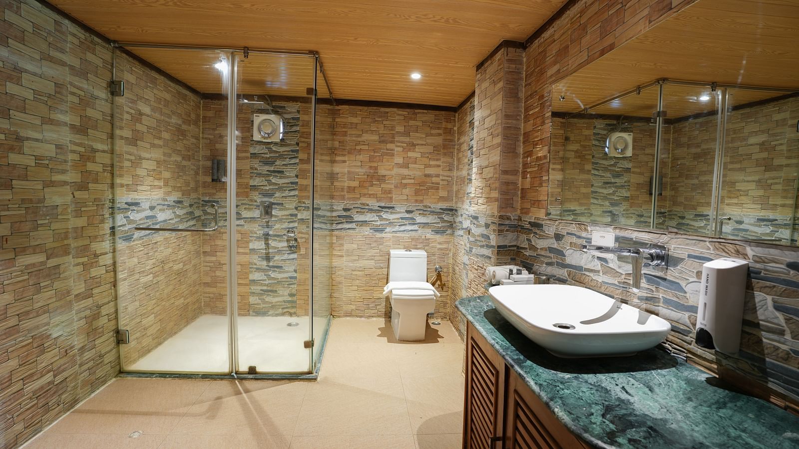 The spacious bathroom of the duplex ethnic villa features glass separating the shower area, a large washbasin with a mirror, and wooden storage space underneath at Summit Green Lake Tea Resort & Spa, Kaziranga