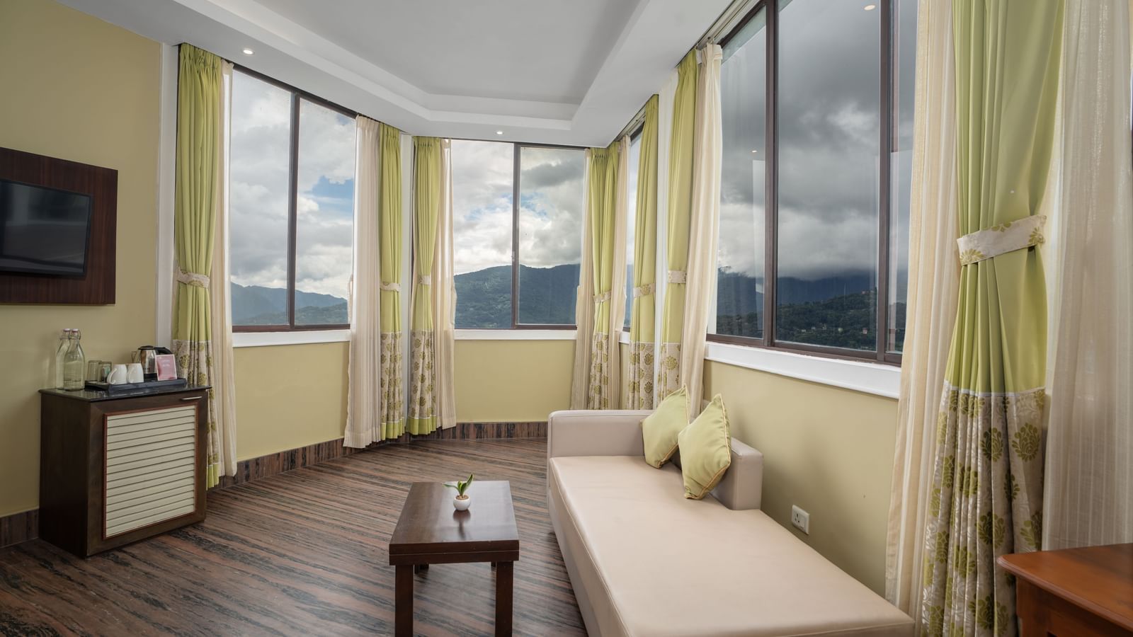 A part of the Honeymoon Suite with Valley View at Summit North Star, Gangtok featuring a sofa with small table, a flat TV screen, a cabinet table with tea and coffee maker, and three windows.
