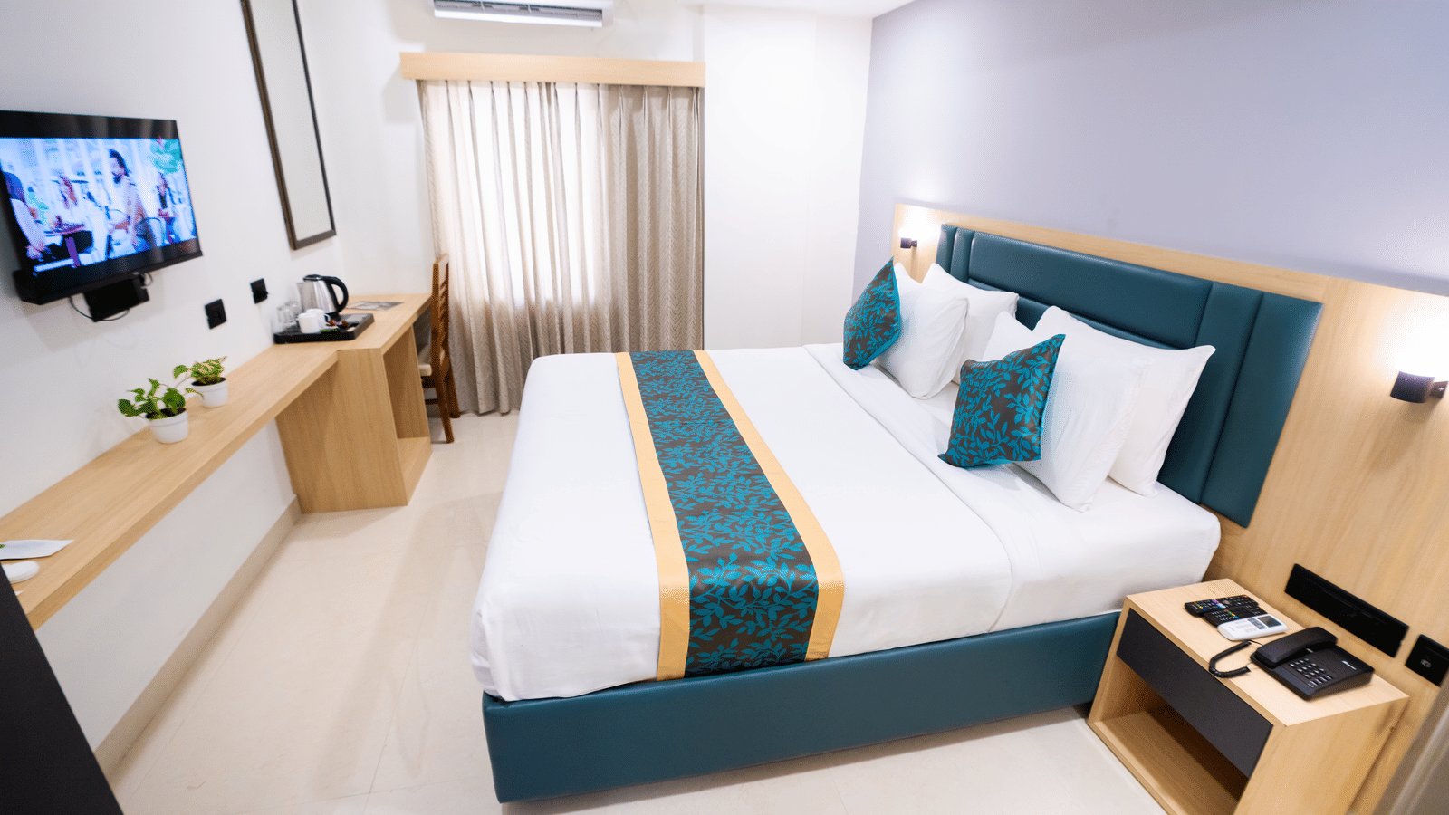 A side view of the room with a large bed, bedside table, wall-mounted TV, and a window with curtains at Sunrise Business Hotel, Hyderabad.