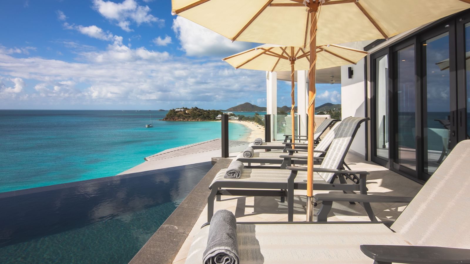 A patio with lounge chairs and umbrellas overlooks a white sand beach and turquoise ocean under a blue sky.