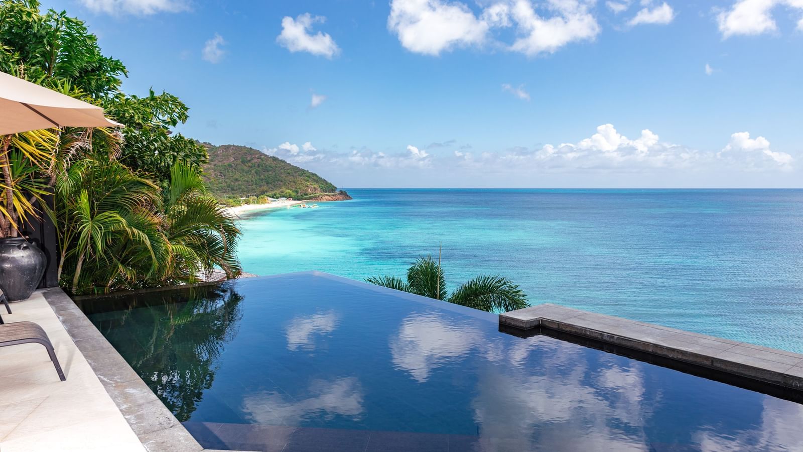An infinity pool that overlooks a turquoise ocean under a bright blue sky, with lush green hills on the left. 