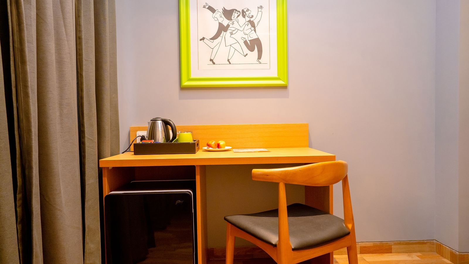 A cosy hotel room workspace with a wooden desk, chair and colorful artwork on the wall - TGI Ameya Grand, Goa