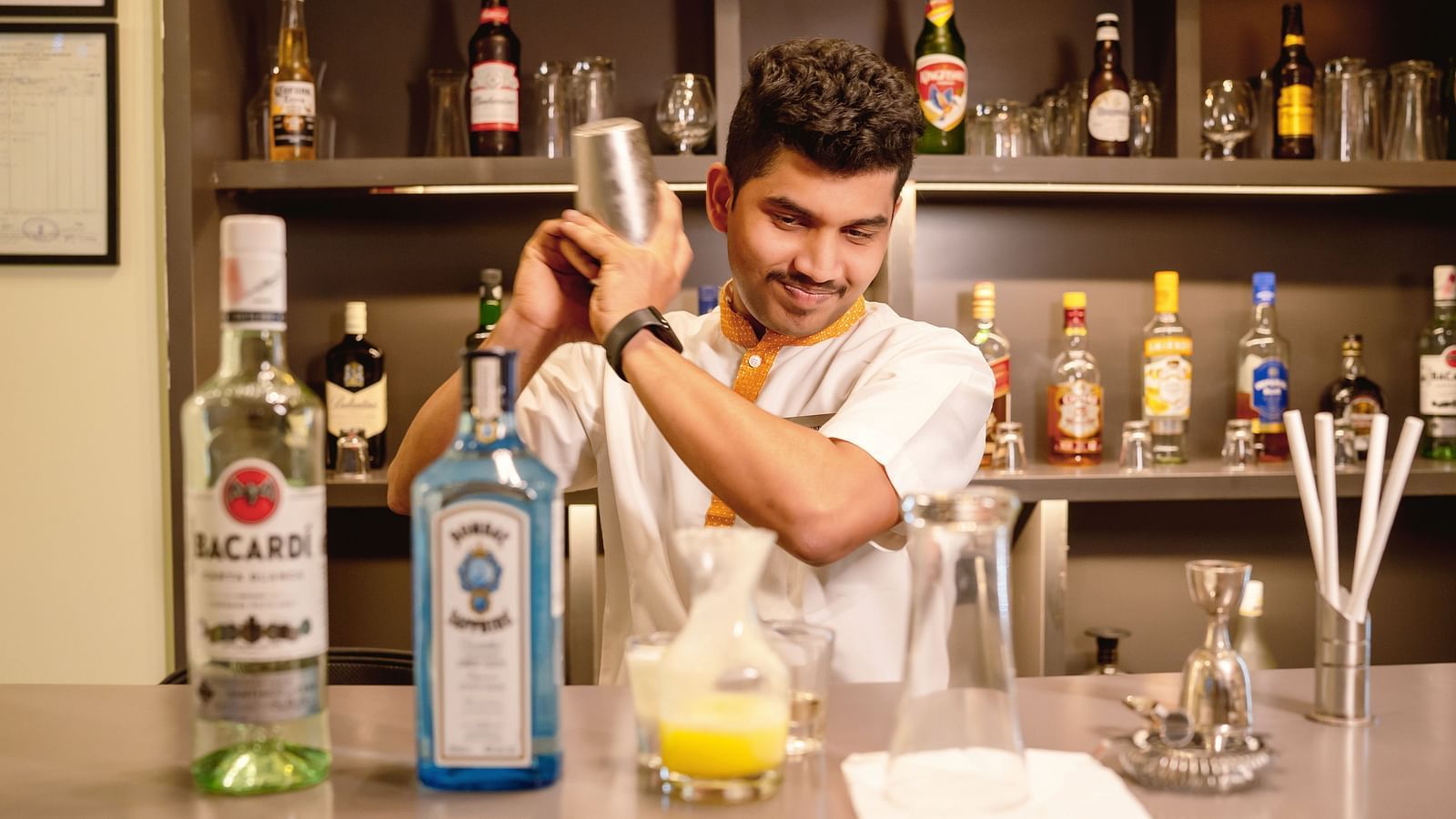 A bartender shaking a cocktail with bottles of alcohol in the background - TGI Ameya Grand, Goa