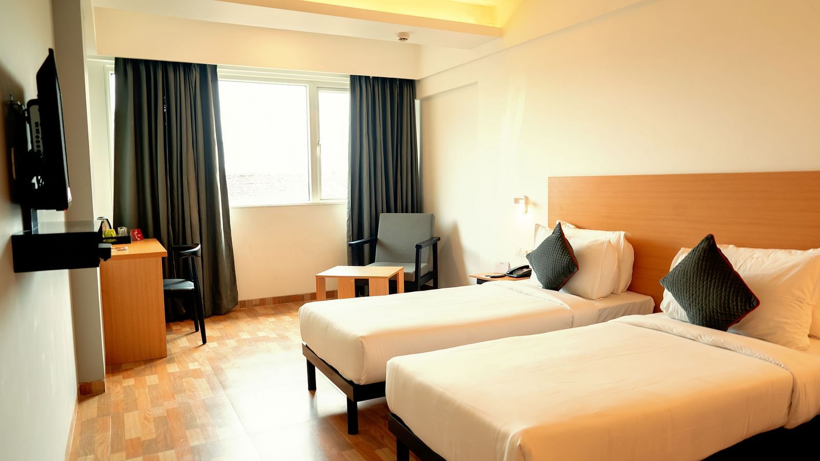 A bright hotel room with two single beds, a desk and a flat-screen TV, with a large window draped in gray curtains - TGI Ameya Grand, Goa