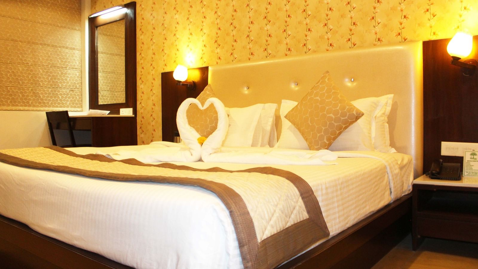 A warmly lit hotel room with a queen size bed, white bedding and a towel art swan on the bedspread - TGI Apple Inn, Jaipur 