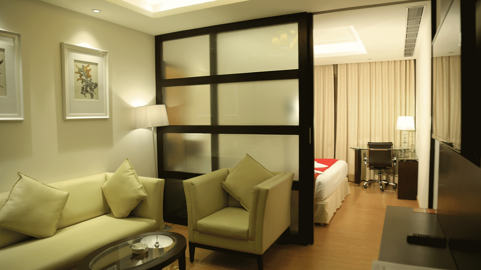 Hotel suite with cream sofa, armchairs and room divider leading to the bed - TGI Grand Fortuna, Hosur