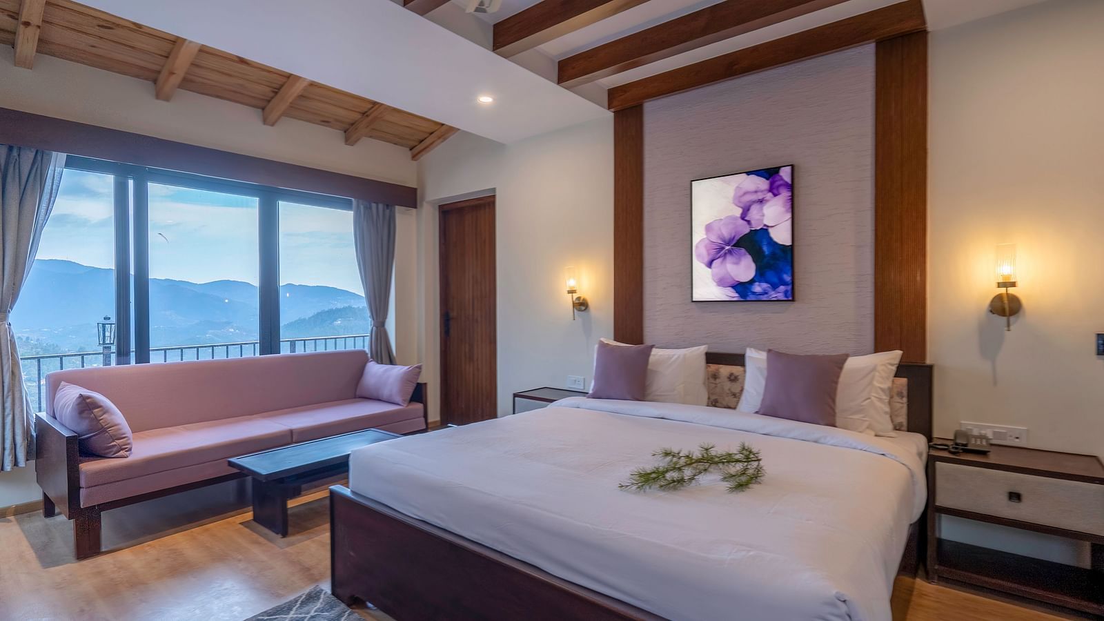 A room with a large bed, a sofa, and a view through a window. Behind the sofa, wall-mounted night lamps are placed on both sides of the bed at The Chamomile Estate.