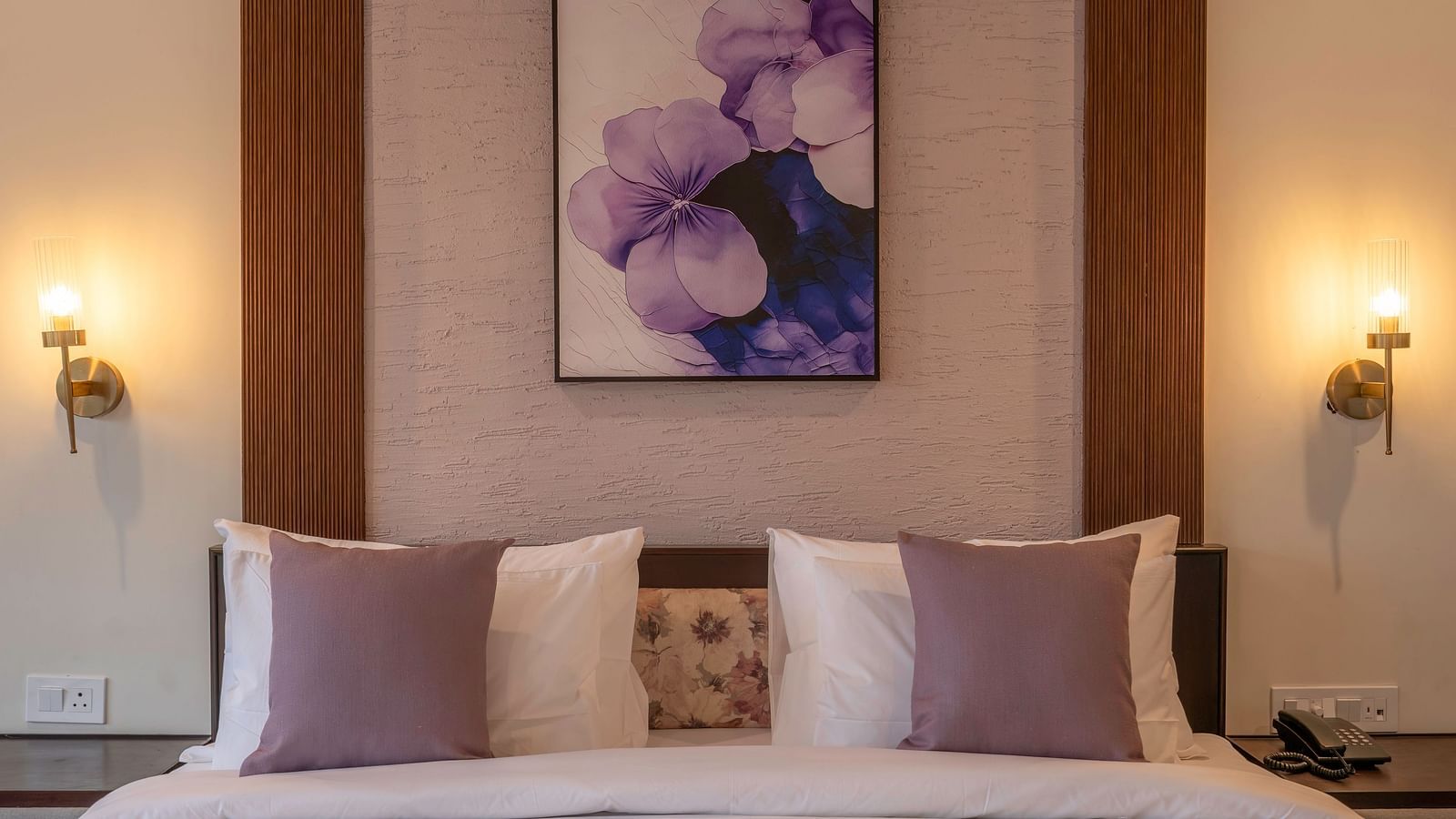 View from the foot of the bed with neatly made sheets and pillows, bedside lamps on both sides, and a floral painting above the headboard at The Chamomile Estate.