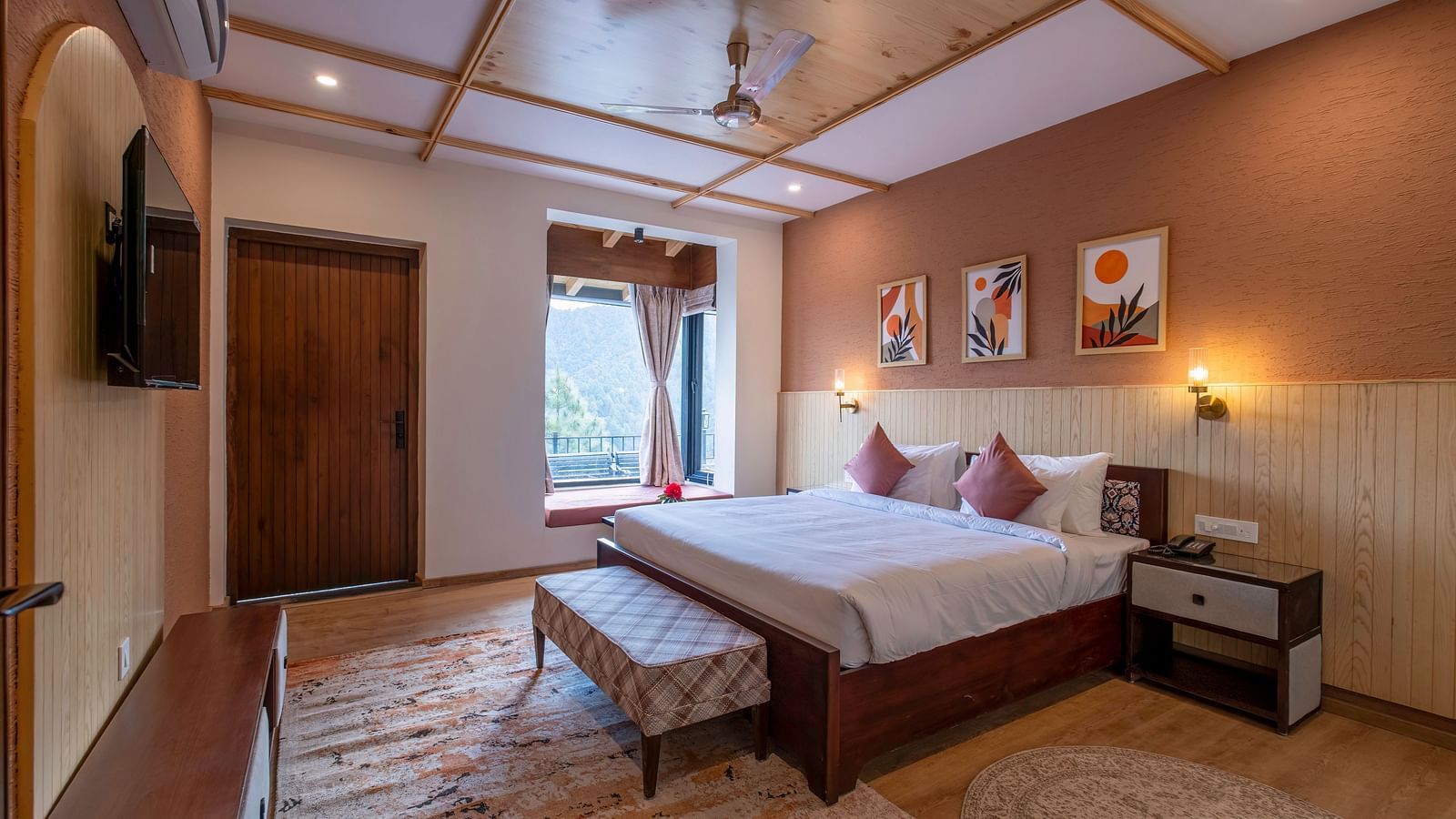 Spacious room with a large bed, bedside table, wall-mounted light above the headboard, a TV with a desk below, and windows and a door to the side at The Chamomile Estate.