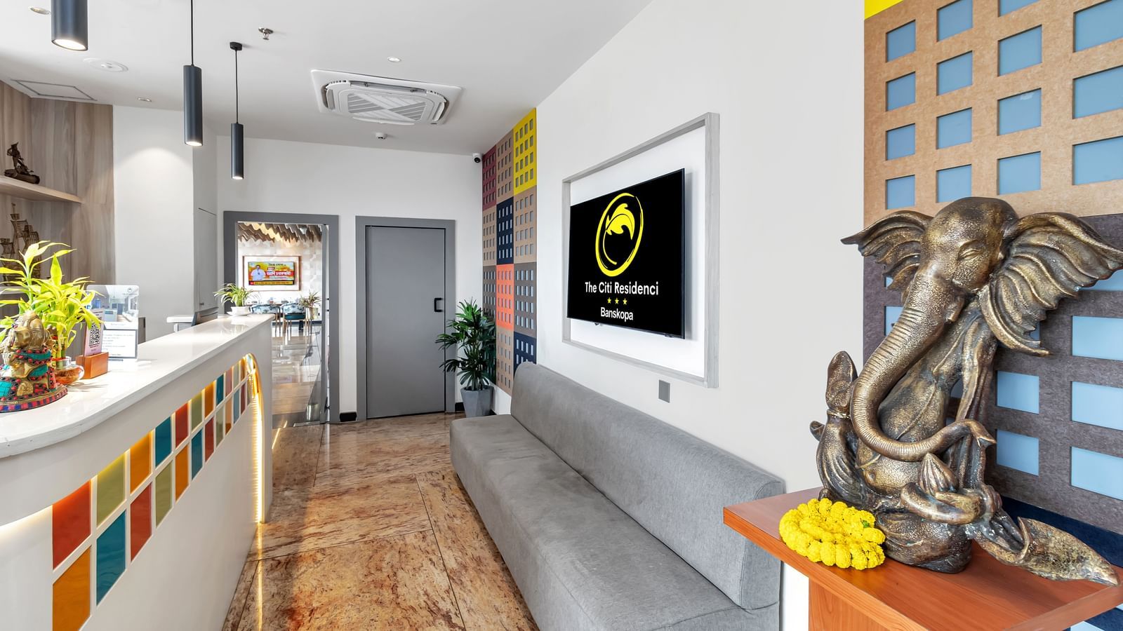 A modern hotel lobby with a colourful reception desk, a grey sofa, a wall-mounted TV, and a Ganesha statue | The Citi Residenci Hotel Banskopa