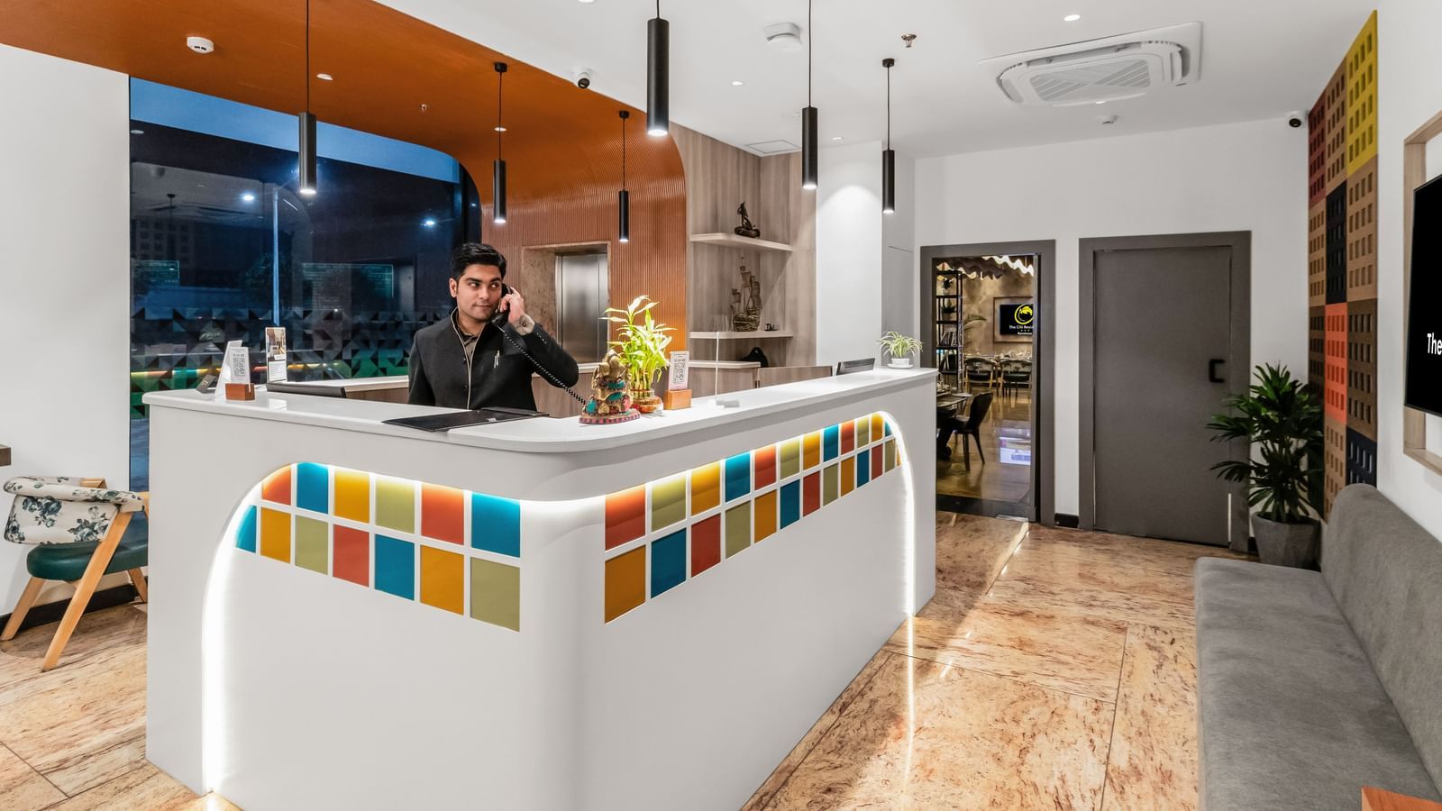 A hotel reception area with a front desk decorated with colourful tiles, a staff member on the phone, pendant lighting, and a seating area | The Citi Residenci Hotel Banskopa