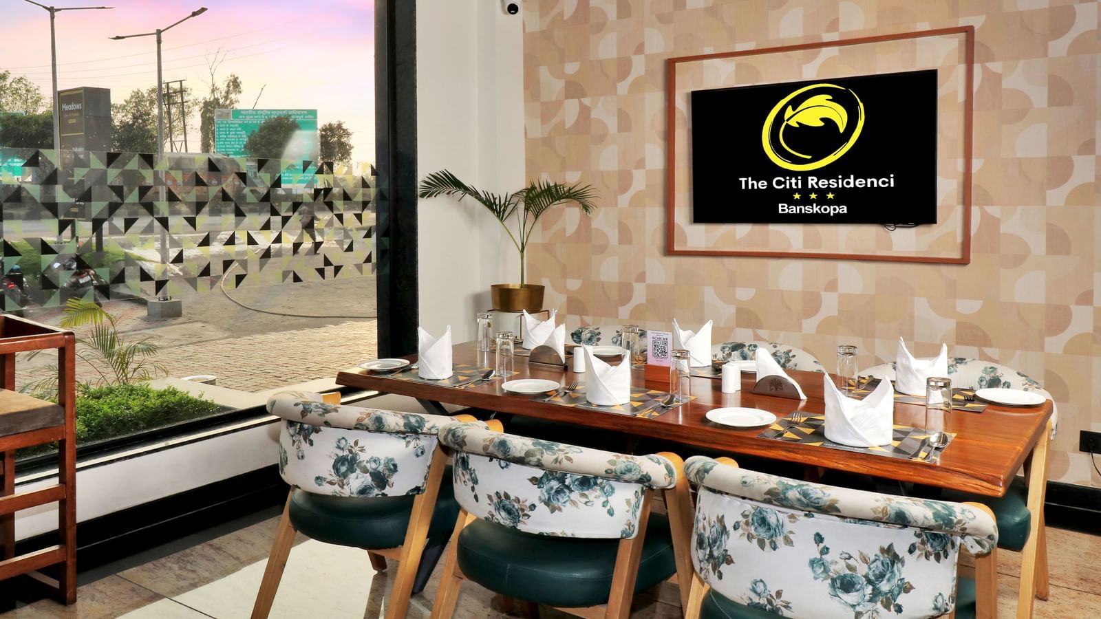 A restaurant dining area with a long wooden table set for guests, surrounded by chairs with floral upholstery, and a framed logo on the textured wall | The Citi Residenci Hotel Banskopa