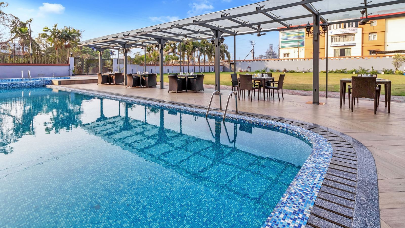 A sparkling blue outdoor swimming pool with mosaic tiling along the edge, surrounded by a paved area and covered dining tables under a clear sky with scattered clouds  | The Citi Residenci Hotel Banskopa