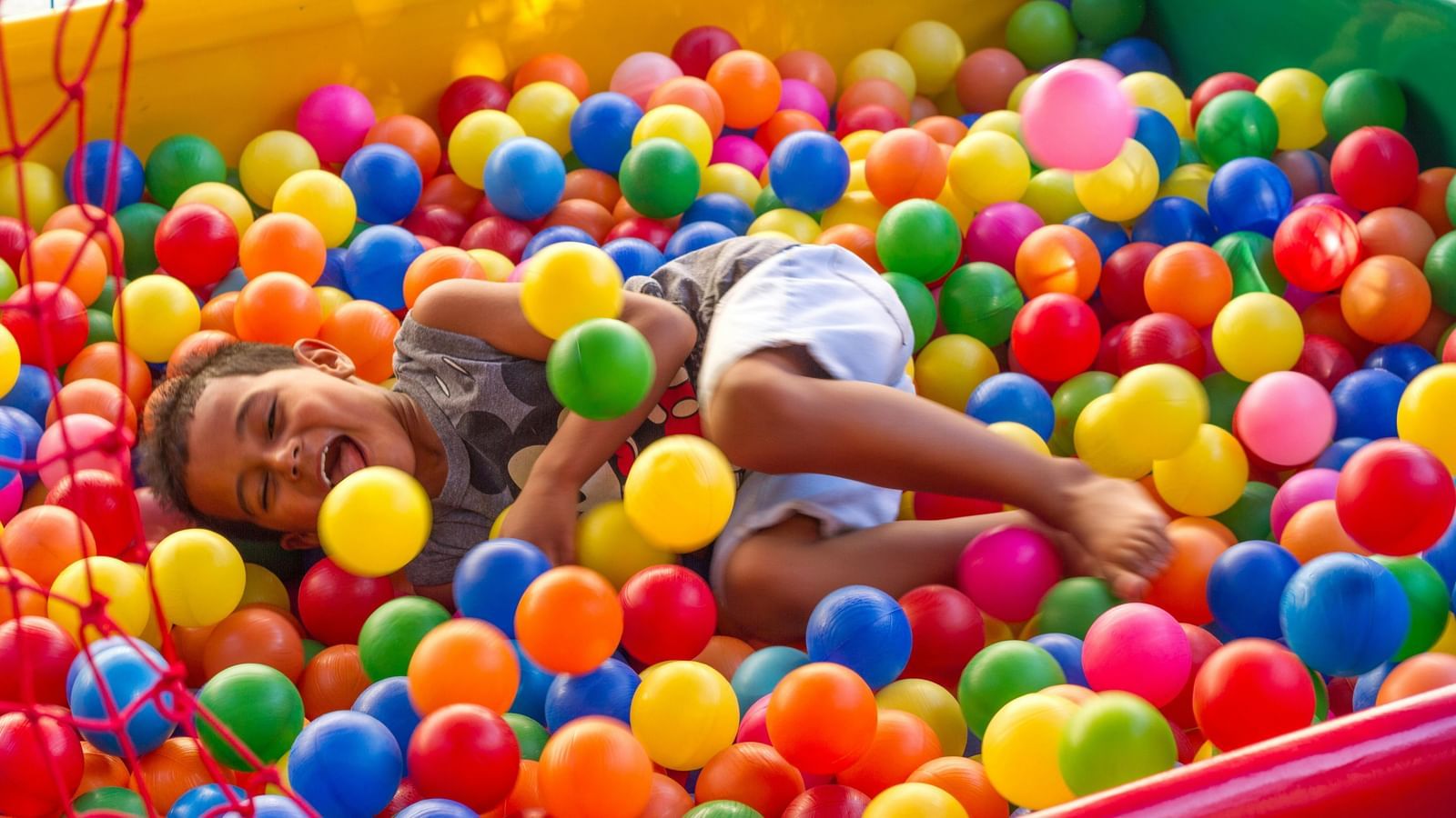 A smiling child lying and playing in a ball pit filled with numerous plastic balls at an indoor play area or playground.