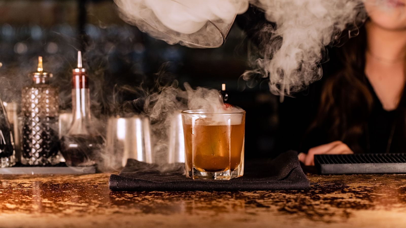 A bartender prepares a drink, with smoke rising from the drink, surrounded by bar tools and ingredients.- The Grand Hotel & Ballroom, McKinney