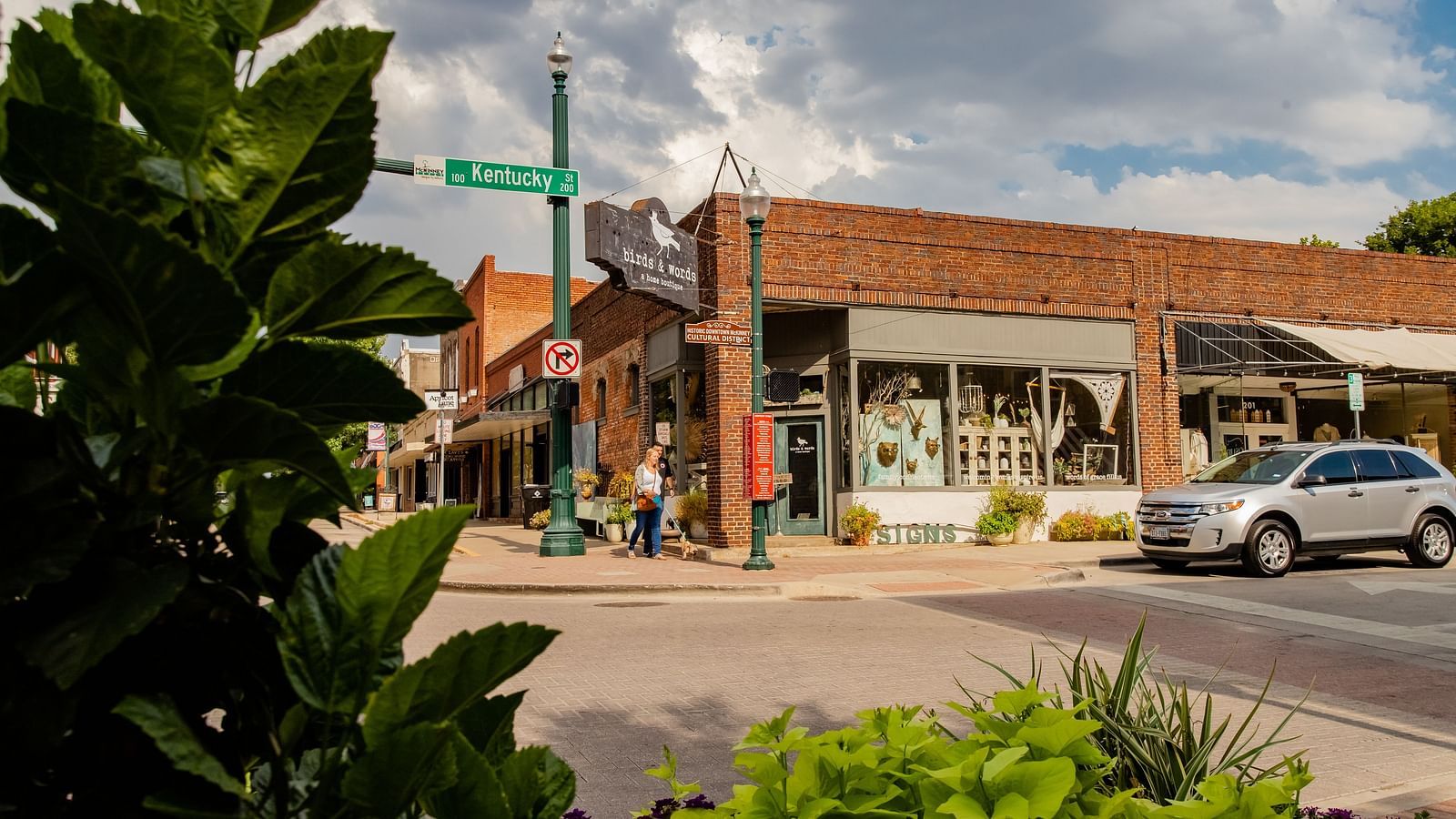 A street lined with shops, trees, a traffic light, and cars parked along the road, viewed from a landscaped corner.- The Grand Hotel & Ballroom, McKinney