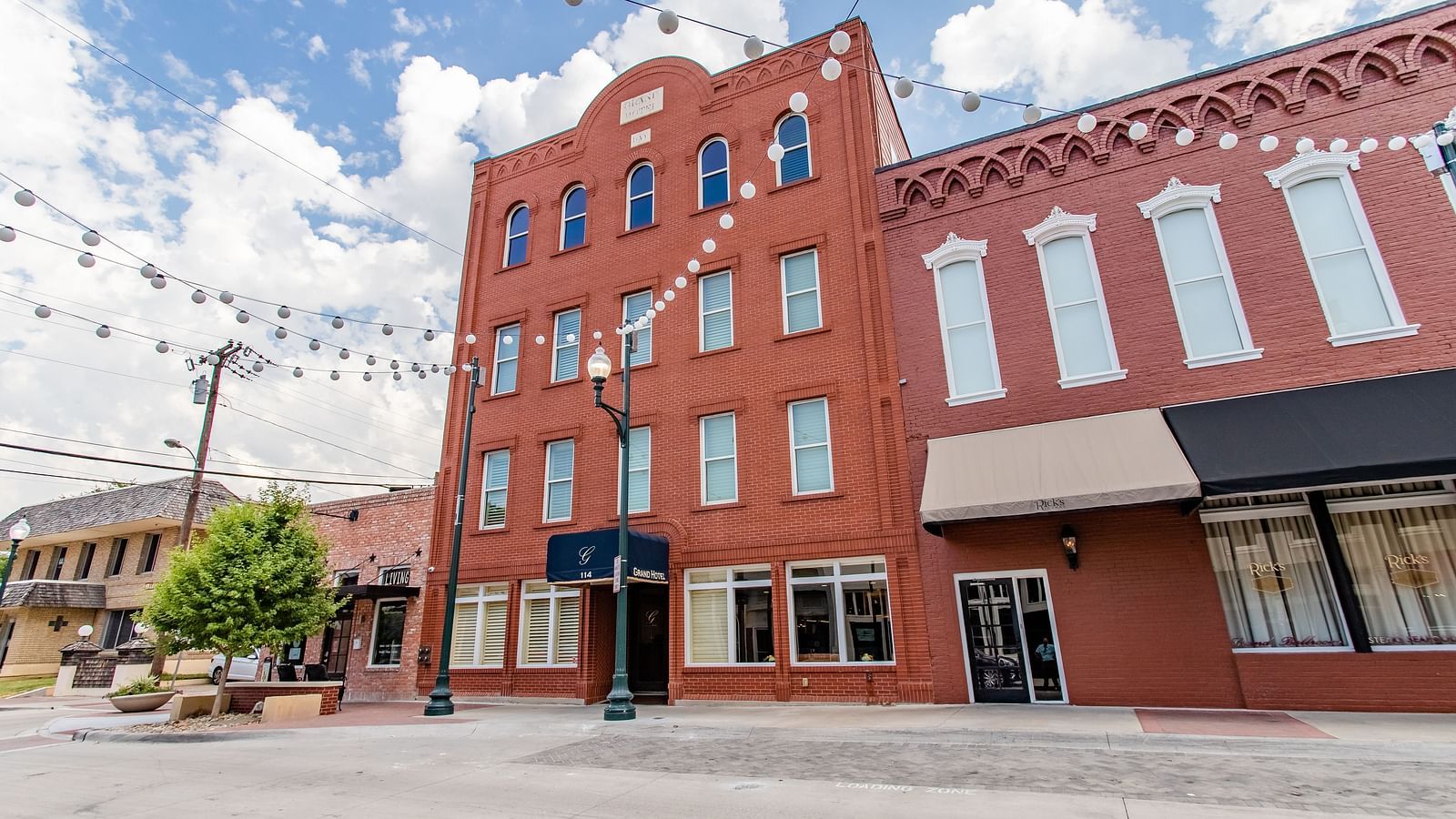 A multi-story brick building with large windows, awnings, and string lights hanging above the street.- The Grand Hotel & Ballroom, McKinney
