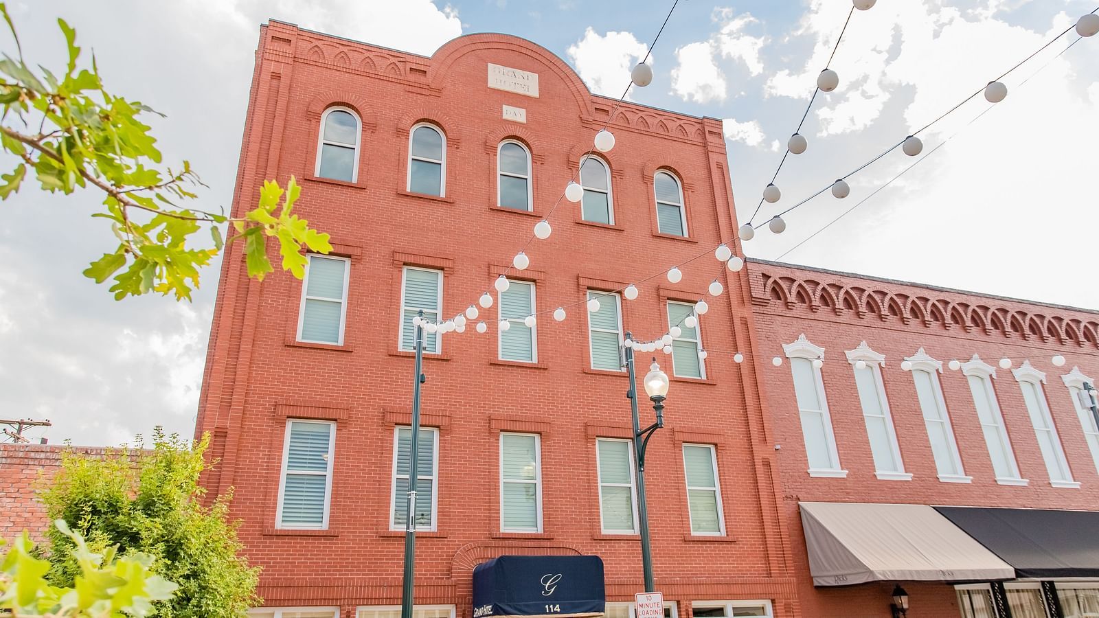 A tall brick building with decorative trim, multiple floors, and a covered entrance with a small awning.- The Grand Hotel & Ballroom, McKinney