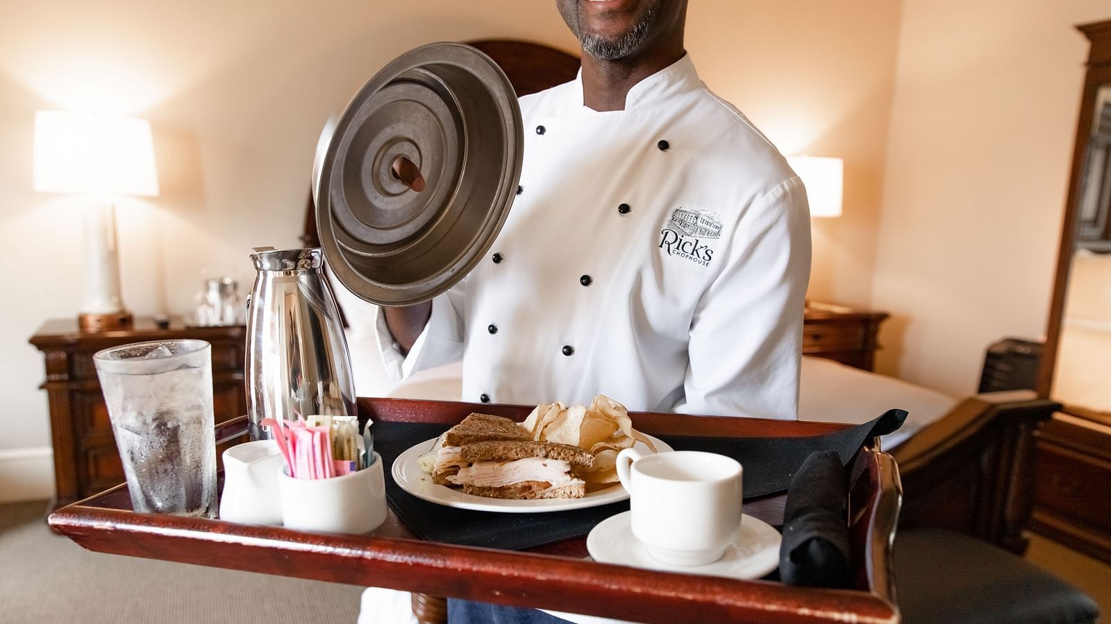 A close-up of a smiling waiter in a chef’s uniform carrying a stray with a covered dish, drink, and condiments.- The Grand Hotel & Ballroom, McKinney
