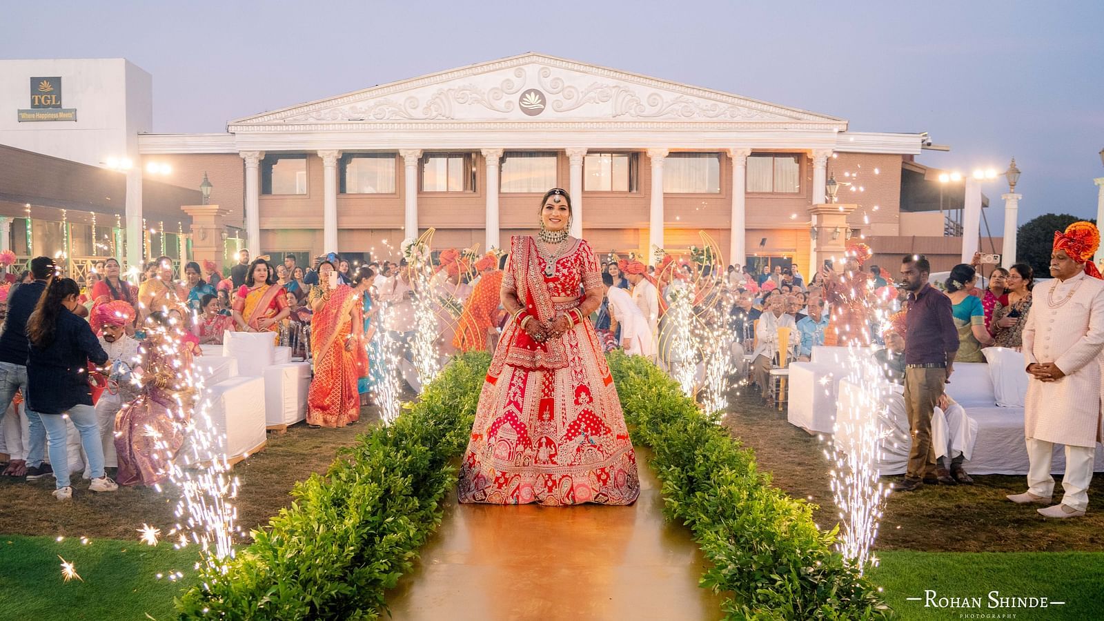 A wedding procession with the bride and groom walking on a flower-lined path, accompanied by people and celebratory sparklers. A large building is in the background - The Grand Legacy Resort and Spa