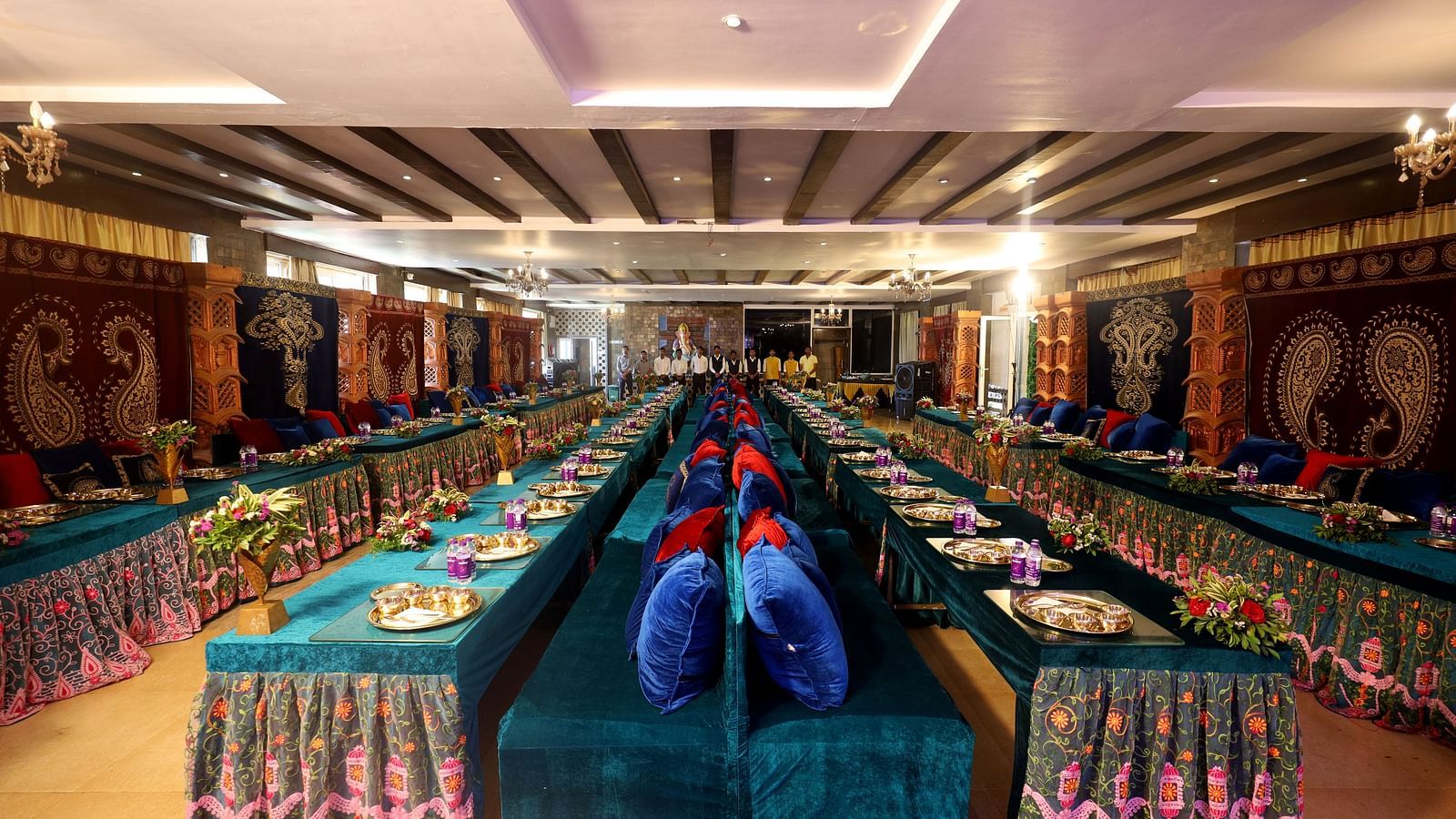 A banquet hall featuring a decorated long table where plates and water bottles are placed and a long chair with cushions paired with it at The Grand Legacy Resort & Spa