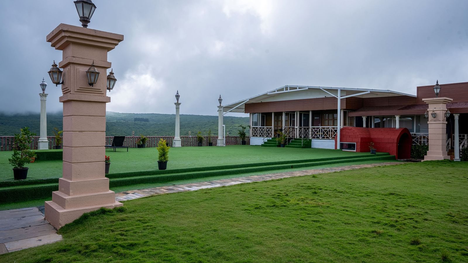 A lush green lawn with a paved pathway leading to a grand building with pillars and arches. The building is surrounded by trees and has a beautiful view of the surrounding landscape.