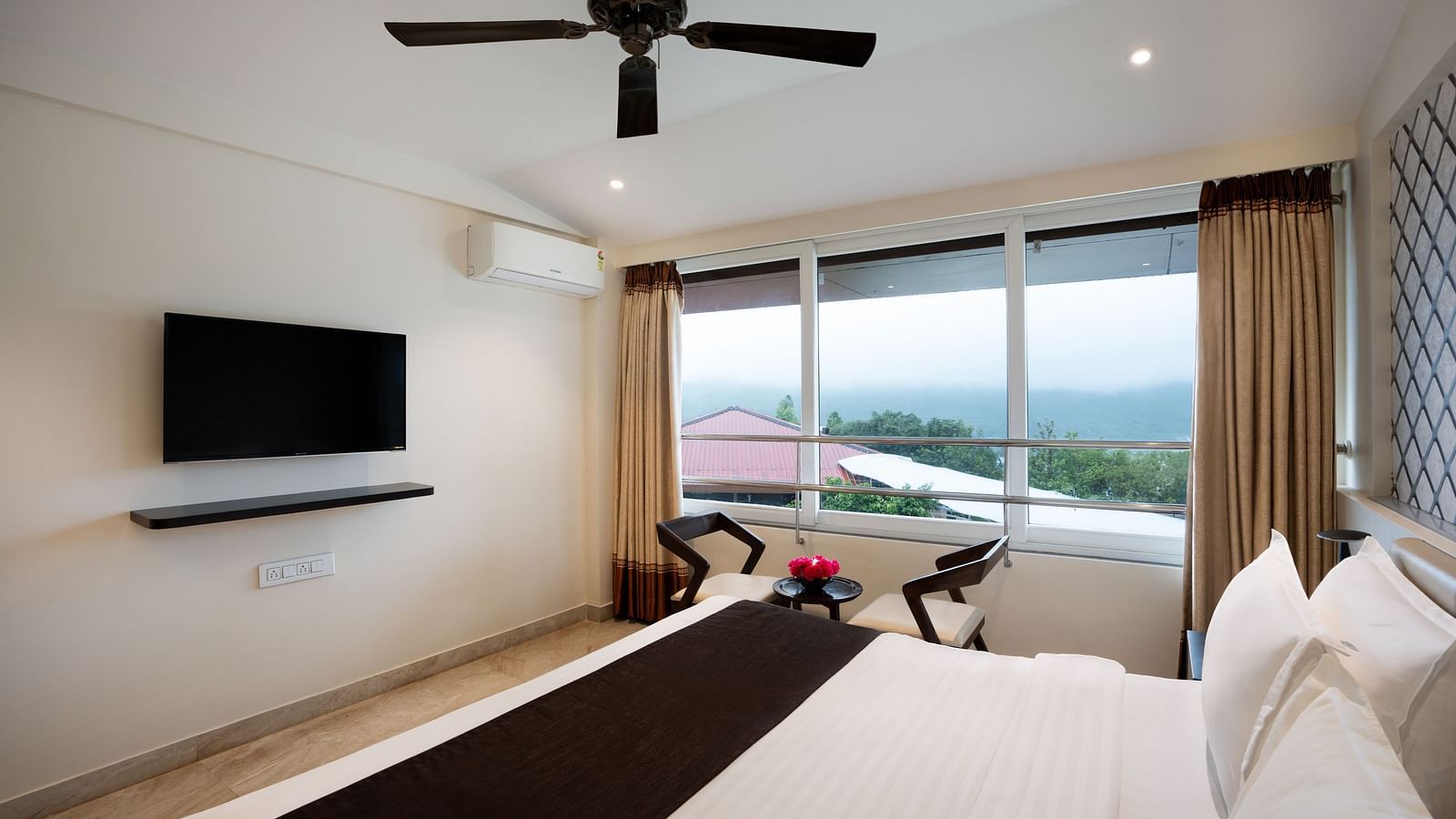 A spacious room with neutral-toned walls, a large bed, a flat-screen TV, and large windows offering natural light.