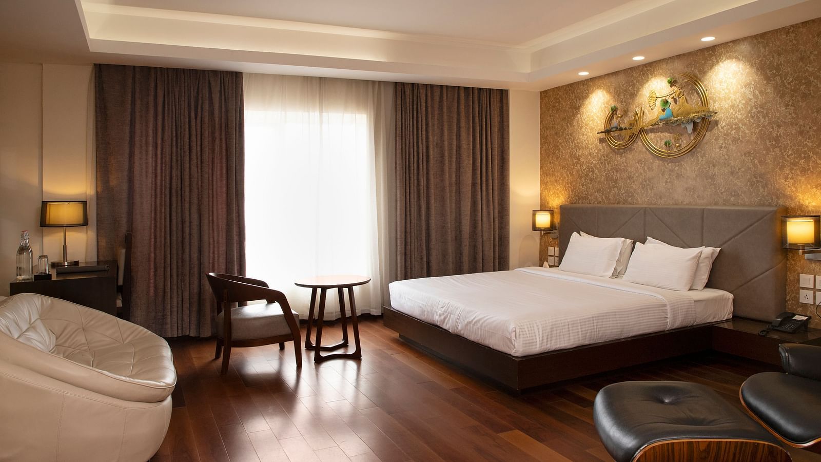 A spacious room featuring a large bed, a cosy seating area, elegant curtains, soft ambient lighting, and expansive windows with curtains drawn to the side at The Hamuse Hotel, Kodaikanal.