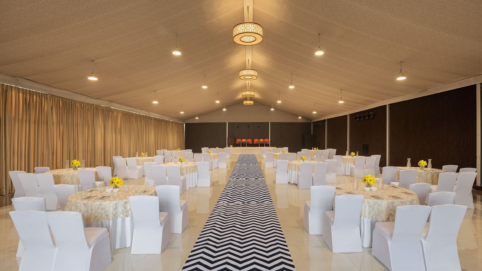 clusters of chairs and tables clad in white covers on either side of a pathway with the chairs facing the other end of the hall - The Hideaway Banquets & Lawns, Greater Noida