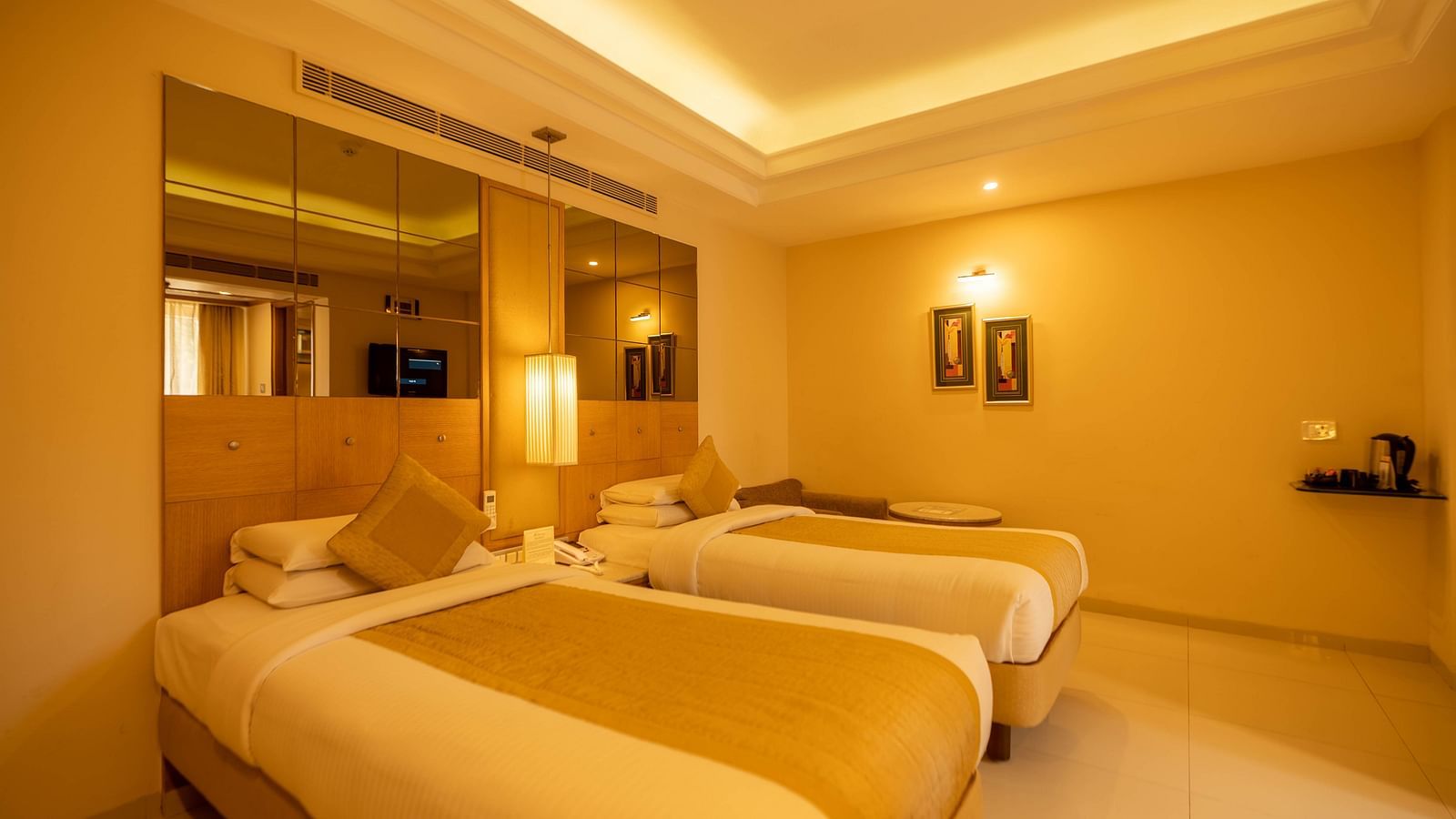 Elegant twin bedroom with golden hues, plush bedding, ambient lighting, and a mirrored wardrobe for a luxurious stay- The President Hotel Jayanagar, Bangalore