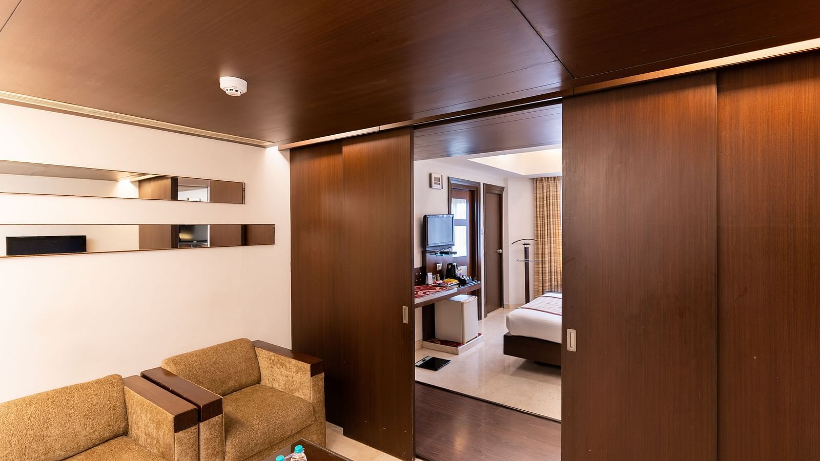 A hotel suite featuring sleek wooden finishes, a cosy sitting area, and a peek into the adjoining bedroom, creating a seamless living space - The President Hotel Jayanagar, Bangalore