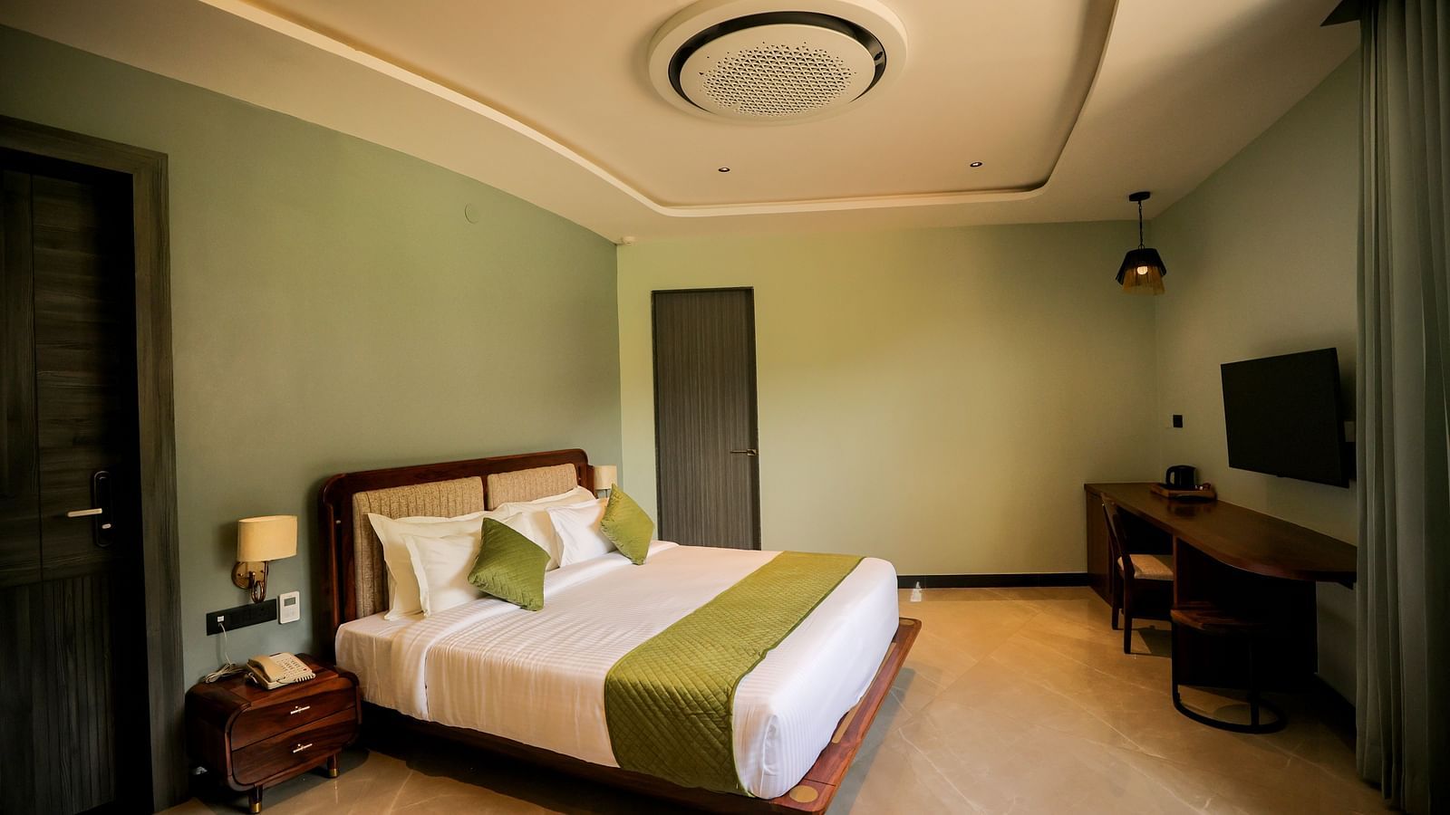 Valley View Room furnished with a king size bed dressed in white linens and a green accent runner, complemented by a wooden side table and a television mounted on the wall at The Qualia Resort Club & Brewery, Udaipur.