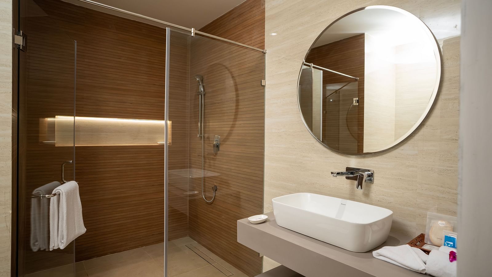 The bathroom featuring a wash basin along with a round mirror above it and a shower area separated by tall glass doors at The Shore, Trishvam.