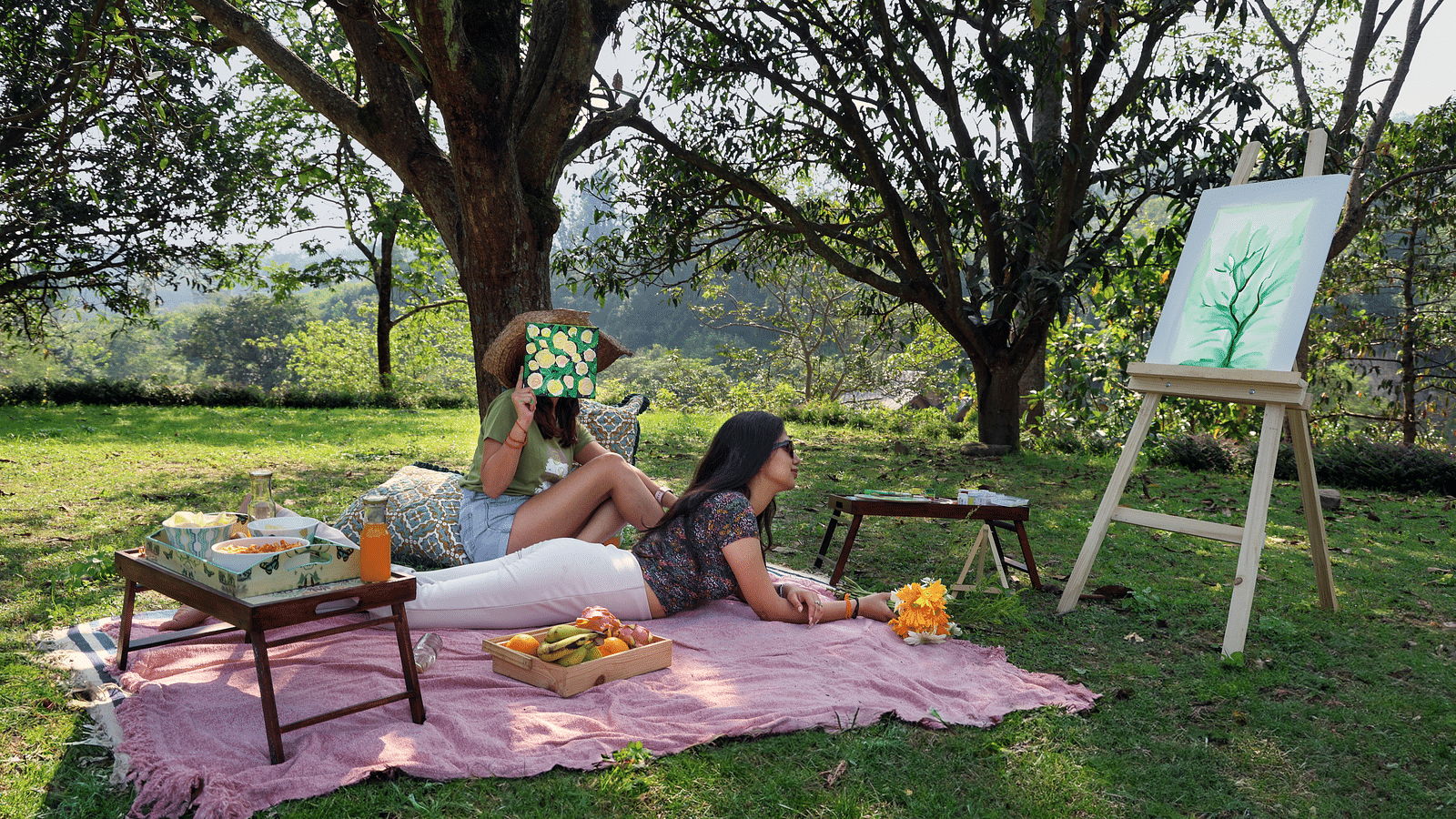 Two people laying on a picnic mat with a snacks table and watching the canvas in a grassfield at the Sylvan Retreat, Dehradun.