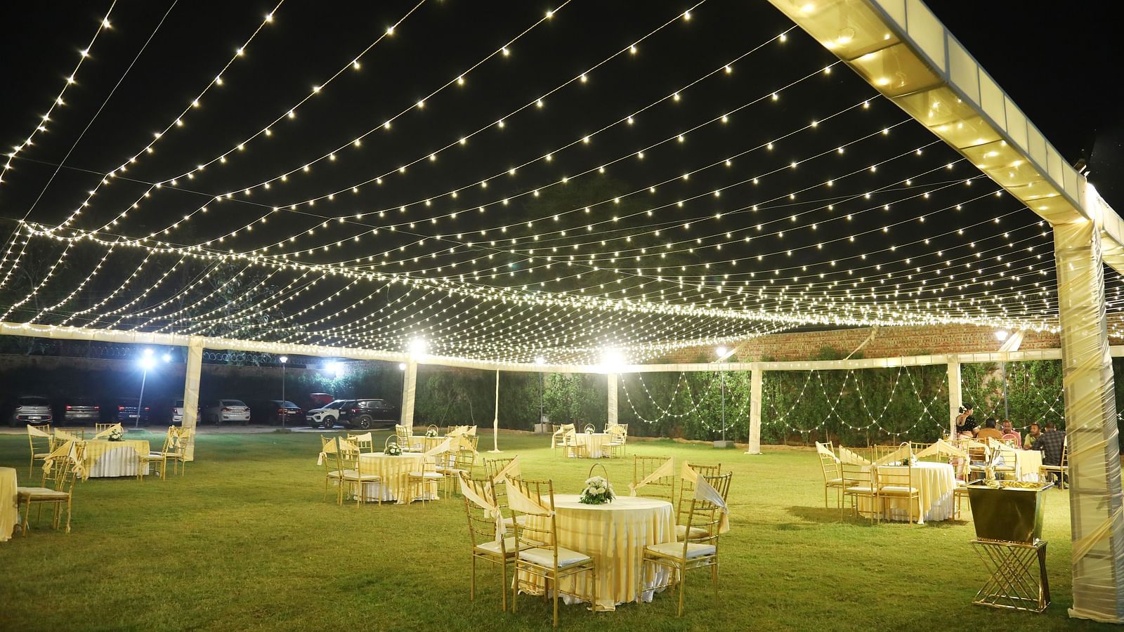 A large lawn at Tisya Farms, Gurgaon with round tables, chairs, and string lights set up for an evening event.