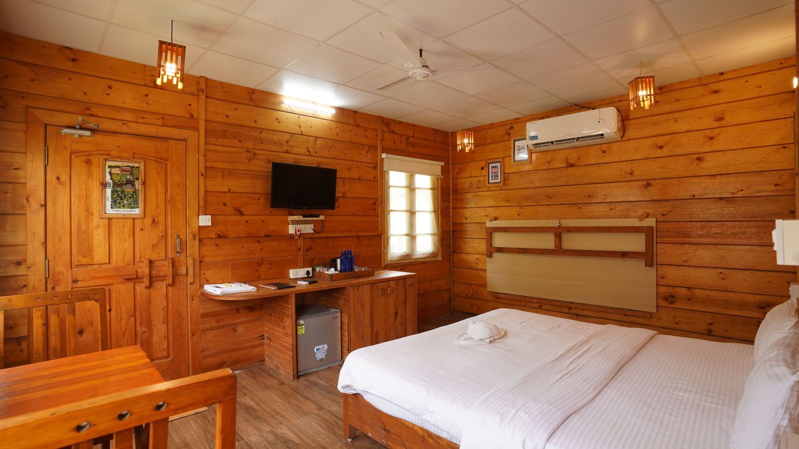 A wooden-clad hotel room featuring a king size bed, desk, television, and small dining area, bathed in warm lighting - Tranquil Beach Resort, Harishareshwar