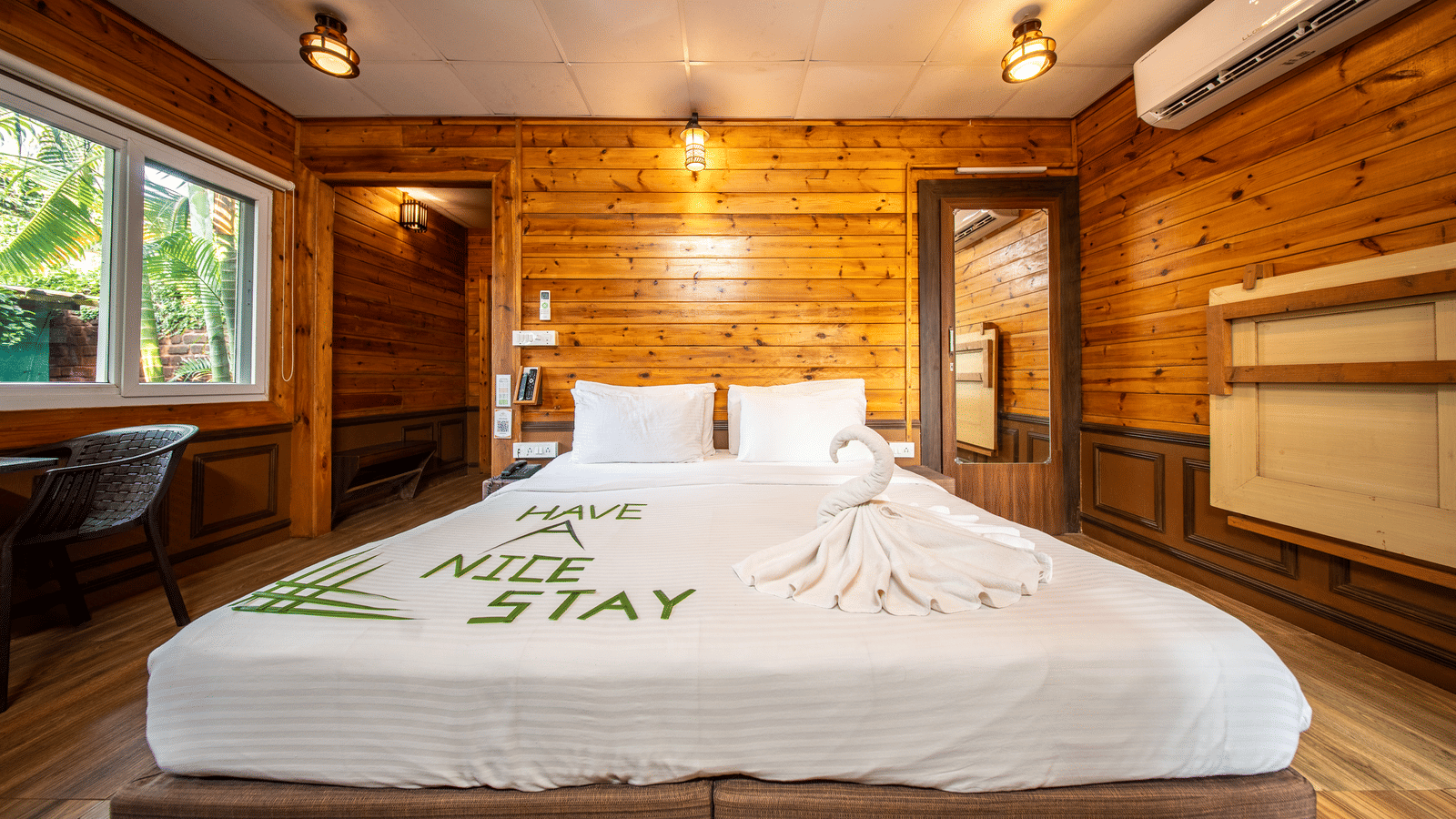 A wooden-furnished room featuring a king-size bed with neatly placed towels, large windows, and Have a Nice Stay written on the bed linen.