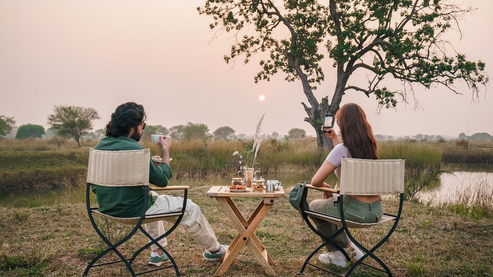 Two people sitting on chairs with a dining setup in an open grassland surrounded by trees at Trees N Tigers, Tadoba.