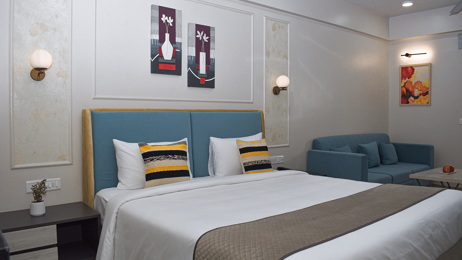 A hotel room with a bed, a blue sofa, a small table, wall-mounted light fixtures, and framed artworks - Urban Suites by BluSalzz, Jaipur