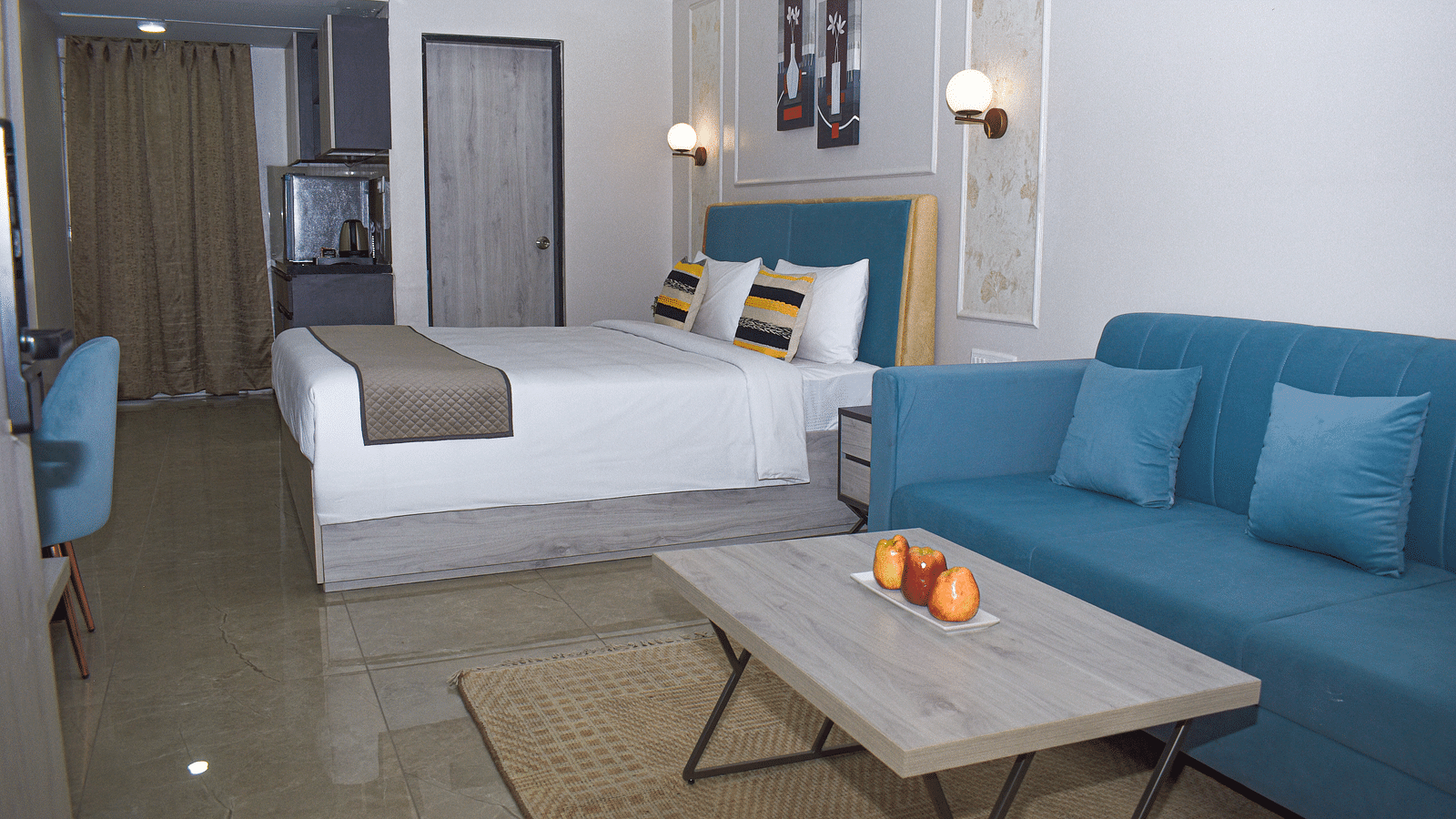 A hotel room with a bed, a blue sofa, a light wood coffee table with fruit, a kitchenette area, and wall-mounted light fixtures - Urban Suites by BluSalzz, Jaipur