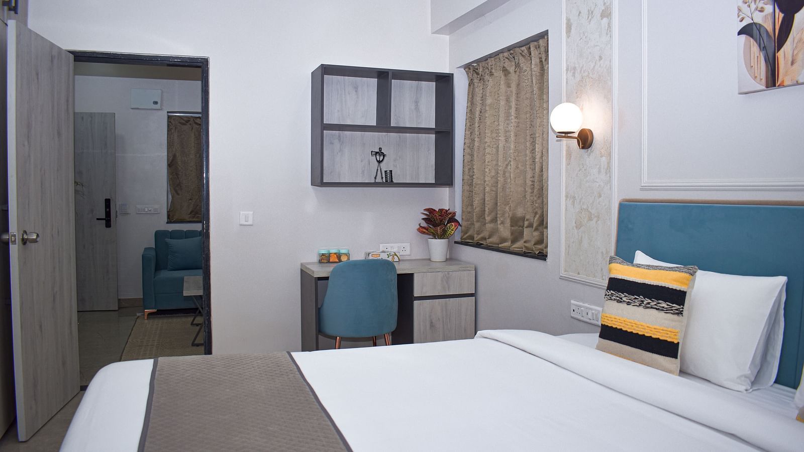 A hotel bedroom with a bed, a desk and chair, a wall cabinet, a window with curtains, and a doorway leading to another room - Urban Suites by BluSalzz, Jaipur
