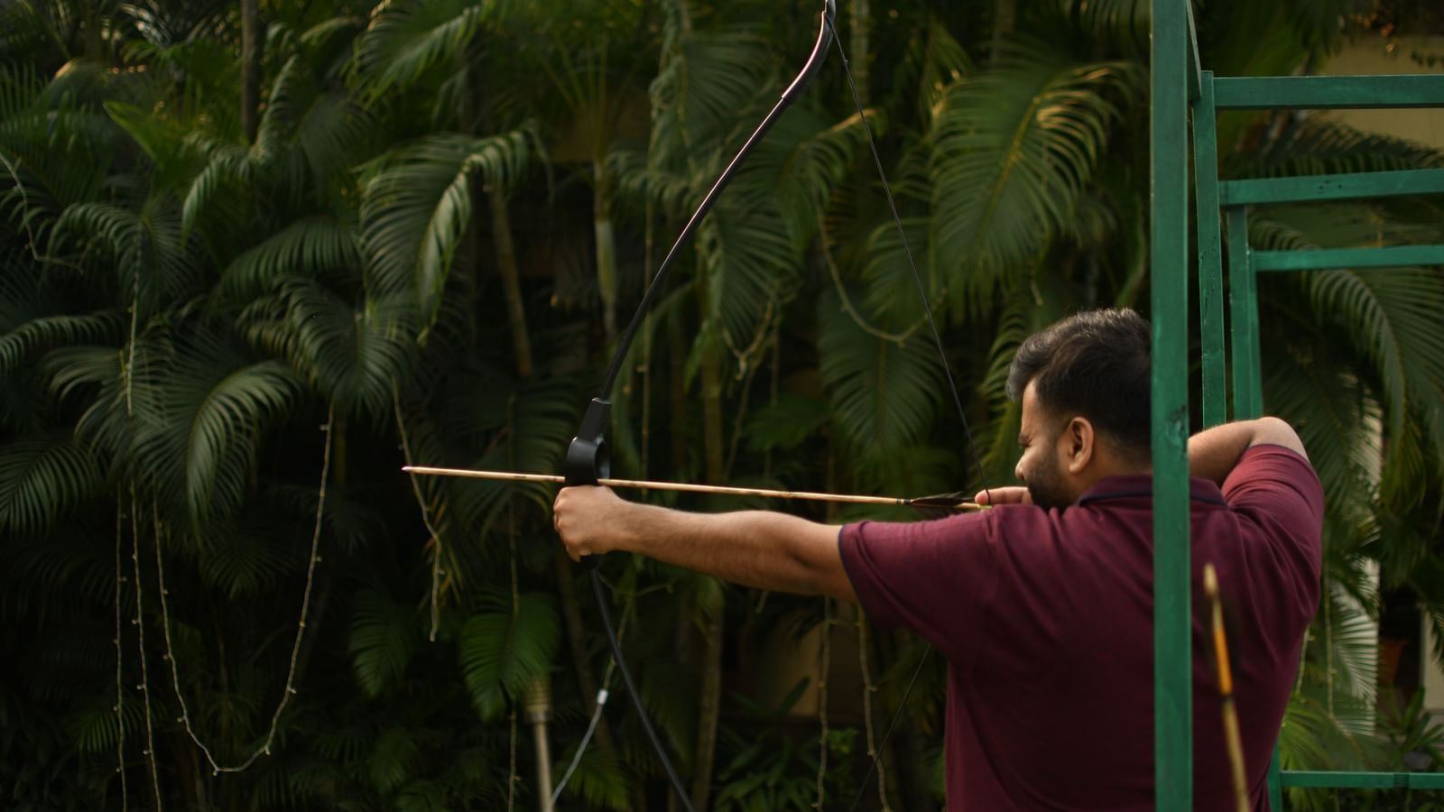 A person aims a long blowgun or similar weapon in a dense, green, natural environment