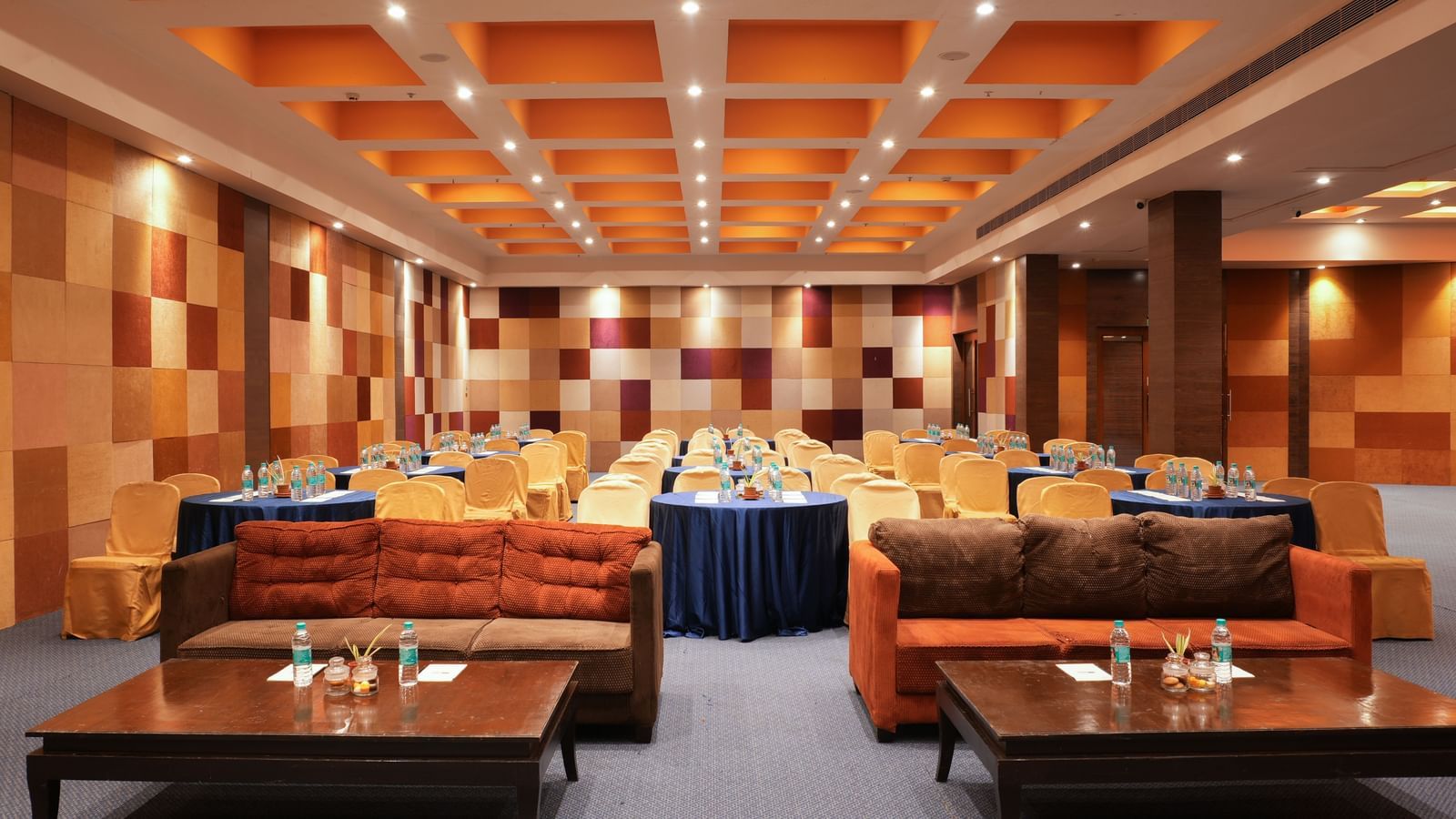 Modern interiors of V-Den at Vedic Village Spa Resort featuring brown plush couches, neatly arranged banquet tables, and elaborative ceilings