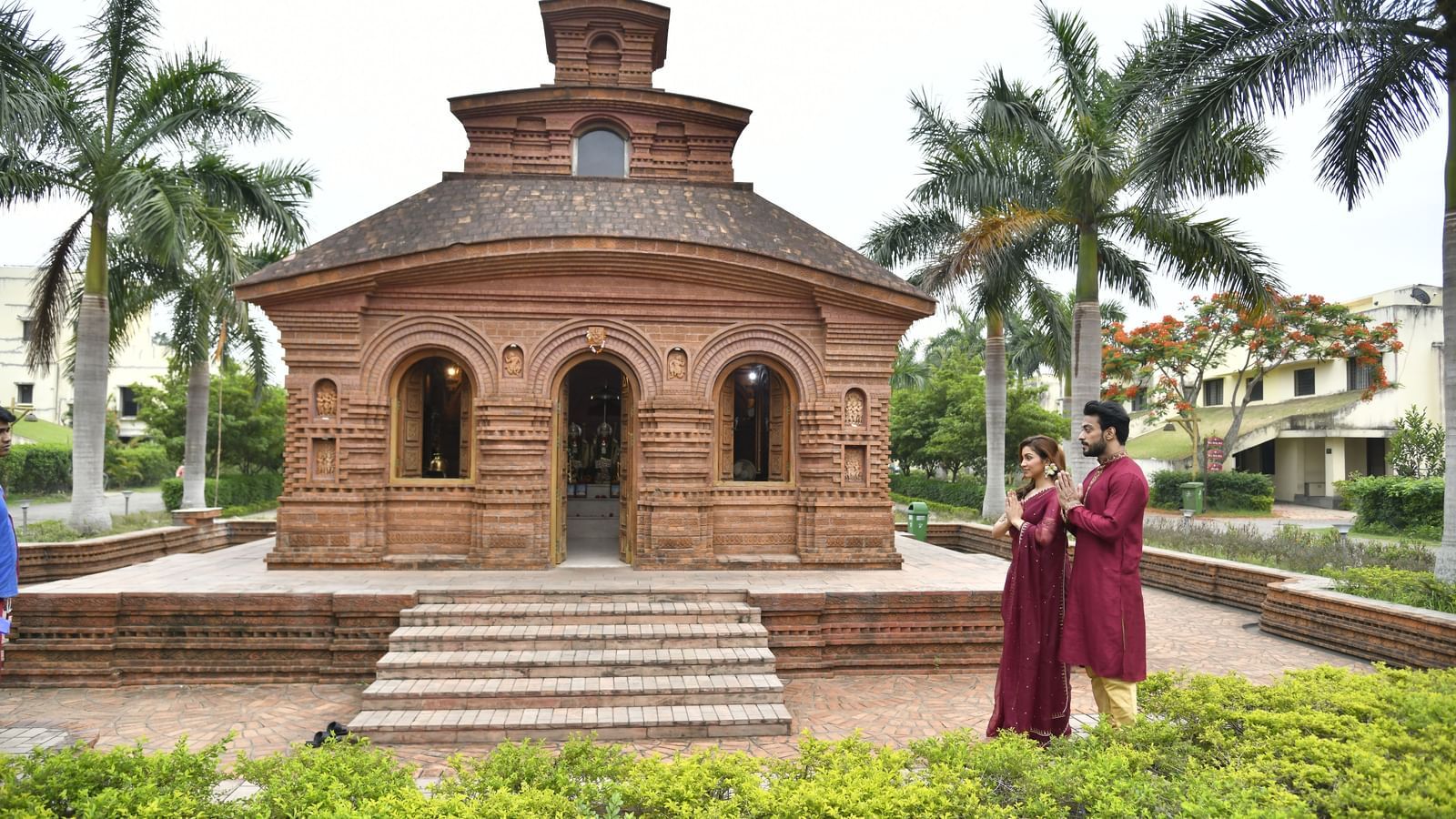 A multi-tiered brick temple-like structure shows stairs leading up to it, with a couple in traditional attire standing at the base and palm trees surrounding it