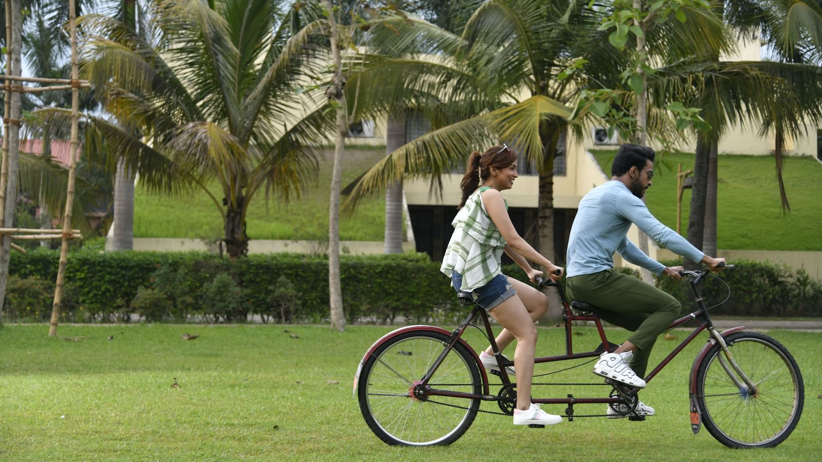A couple rides a tandem bicycle on a grassy lawn with palm trees in the background