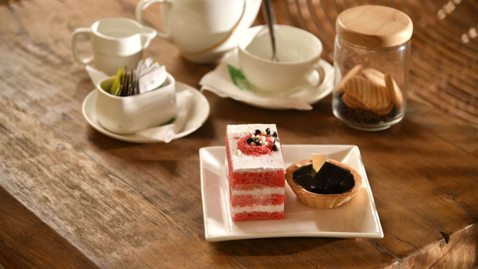 A tea set with cups, saucers, and a teapot is arranged on a table alongside a dessert | Chai