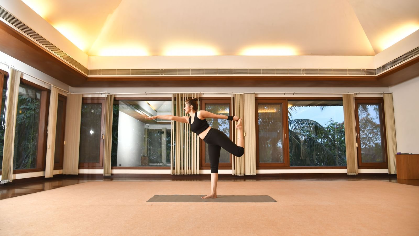 A woman in black activewear practices a balancing yoga pose on one leg in a spacious room with wooden floors, large windows, and mirrored walls