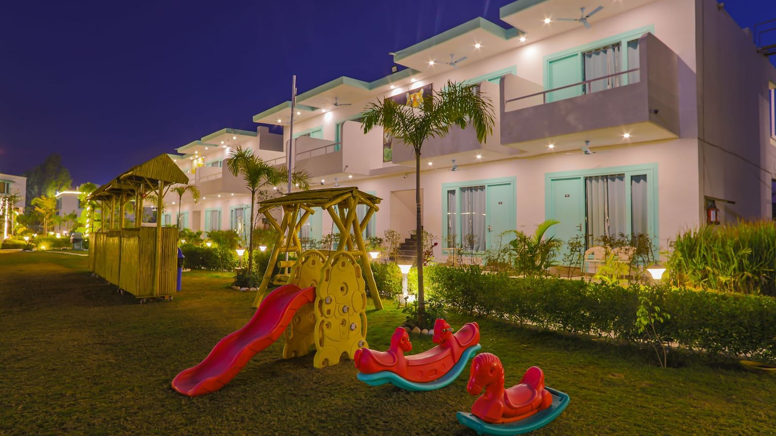 Children’s play area with bright red swings and slides in front of Vedikant Resorts The Mallard Corbett's rooms during evening hours.