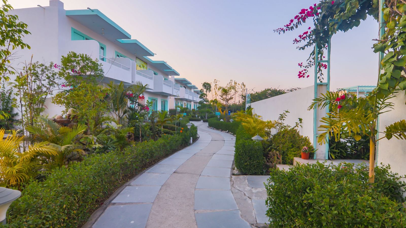 A walkway with flowering plants leads past buildings at Vedikant Resorts The Mallard Corbett at dusk.