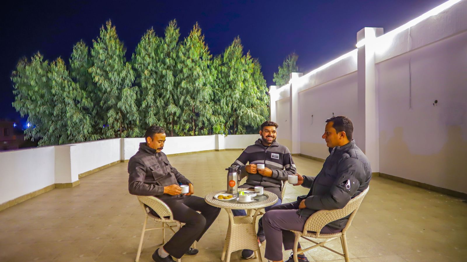 People sit at a table on a rooftop area at Vedikant Resorts The Mallard Corbett at night, surrounded by greenery.